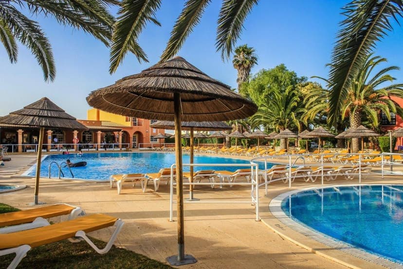 Zwembad met rieten parasols, gele ligbedden en palmbomen bij Colina Village in Carvoeiro