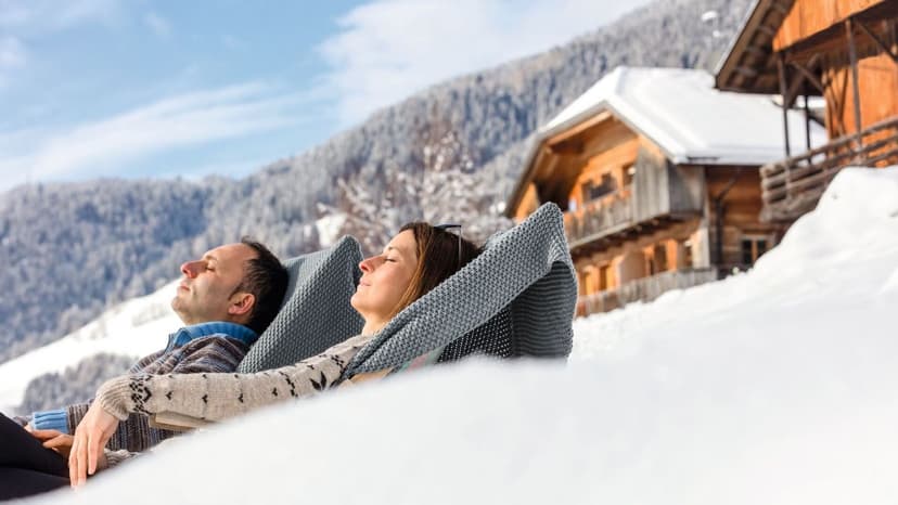 Een koppel ontspant in ligstoelen in de sneeuw voor een houten berghoeve in Zuid-Tirol