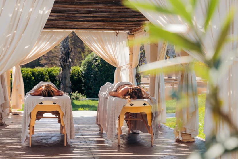 Twee massagetafels onder een open paviljoen met witte gordijnen in een groene hoteltuin op Malta