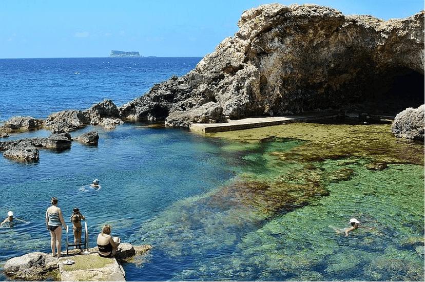 Zwemmers genieten van een natuurlijk rotszwembad met een rotsboog en uitzicht op Comino